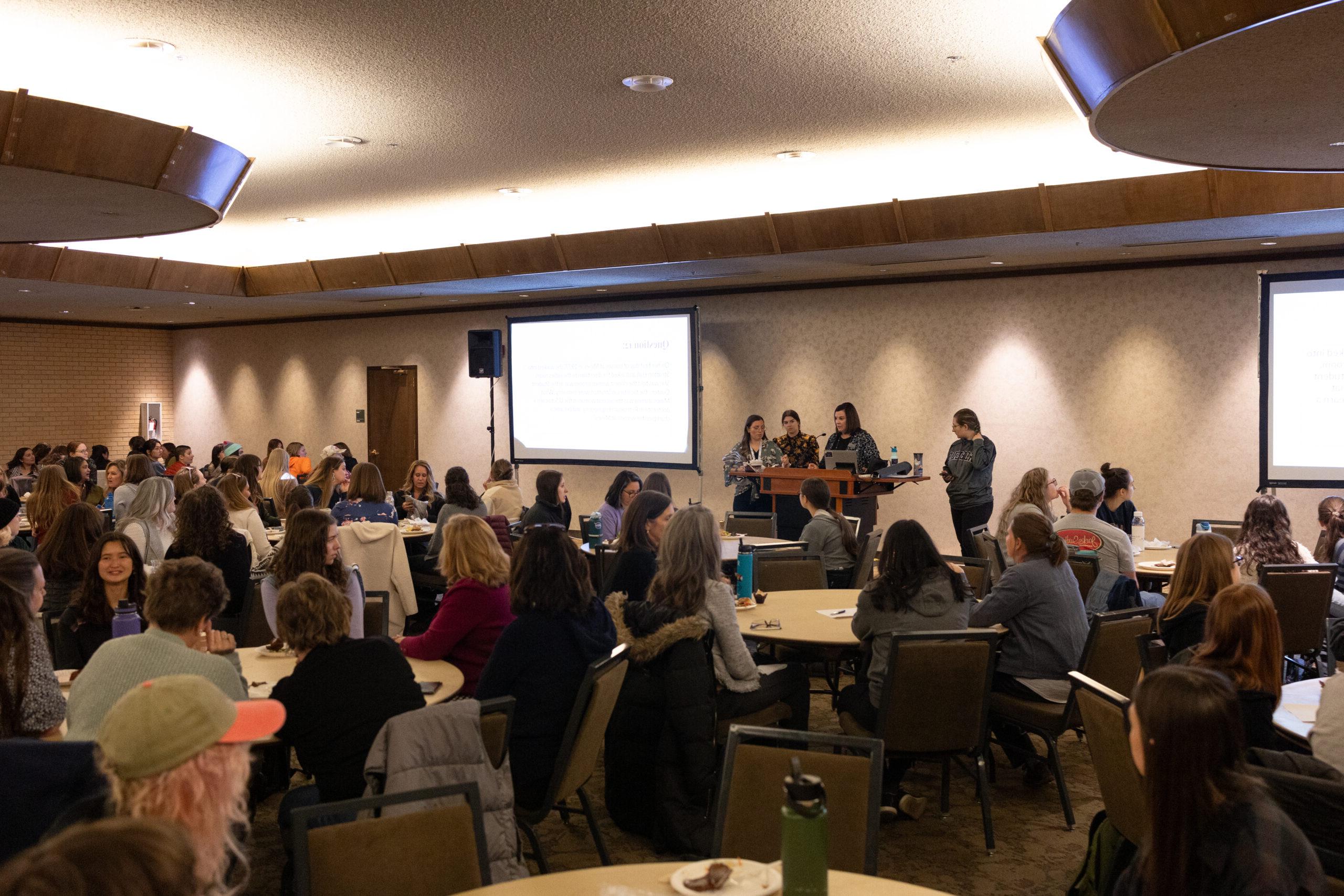 Women sitting at tables looking at screen in a conference room, playing trivia, eating lunch